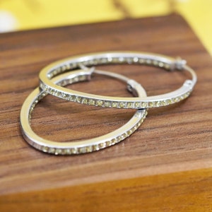 Puede incluir: Un par de pendientes de aro plateados con una fila de pequeñas piedras preciosas amarillas en el borde exterior. Los pendientes están sobre una superficie de madera, con un fondo amarillo.