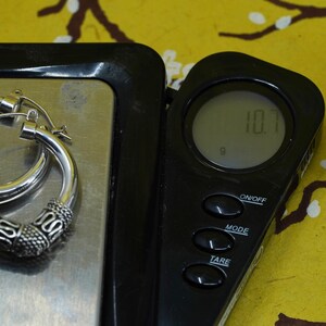 May include: A pair of silver hoop earrings with an ornate design, resting on a digital scale. The scale displays a weight of 10.7 grams. The earrings have a detailed pattern on the lower portion of the hoops.