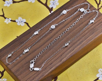 May include: A delicate silver necklace with multiple clear, round gemstones. The necklace is displayed on a wooden surface against a yellow background with a floral design. The necklace has a lobster clasp.