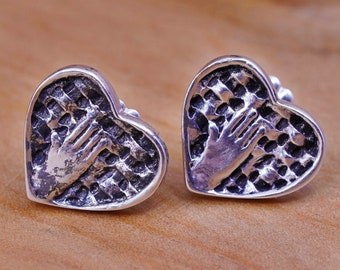 May include: A pair of silver heart-shaped stud earrings. Each earring features a textured surface with a hand design in the center. The earrings are displayed on a wooden surface.