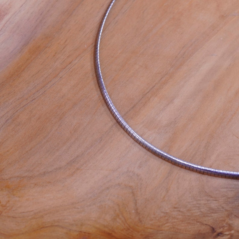 May include: A silver-colored necklace with a smooth, rounded design. The necklace is made of small, connected segments, creating a flexible, sleek appearance. The jewelry is displayed on a light brown wooden surface.