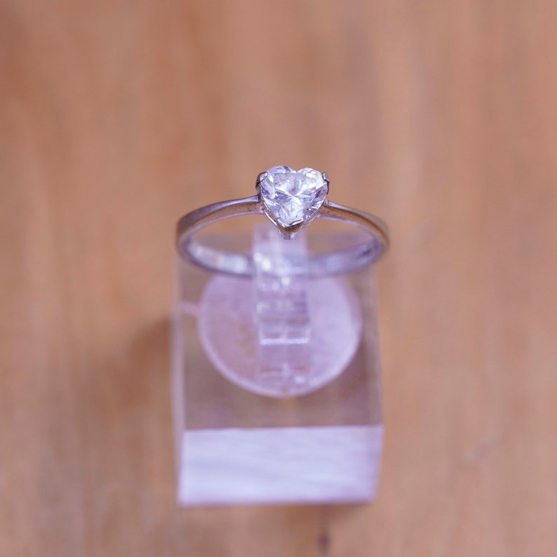 May include: A silver ring with a heart-shaped, clear gemstone. The ring is displayed on a clear, rectangular stand against a light brown background. The gemstone is the focal point, catching the light and reflecting a brilliant sparkle.