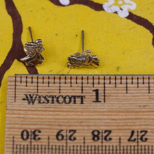 K&ouml;nnte beinhalten: Zwei silberfarbene Ohrstecker, jeweils mit einem detaillierten Design einer Figur mit flie&szlig;endem Haar. Die Ohrringe werden auf einer gelben Oberfl&auml;che mit einem Holzlineal gezeigt, das Messungen anzeigt.