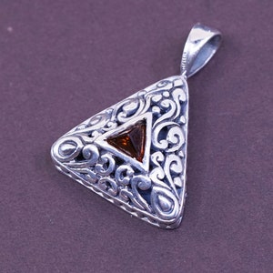 May include: Silver-toned pendant with a triangular shape and intricate scrollwork. A brown, triangular gemstone is set in the center. The pendant has a loop for a chain and is displayed against a dark background.