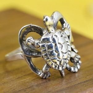 May include: A silver-toned ring featuring a detailed turtle design. The turtle's shell and limbs are intricately crafted, with the turtle's head and feet extending from the ring's band. The ring is set against a wooden surface with a yellow background.