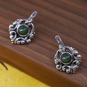 May include: A pair of silver-toned earrings with a green gemstone center. The earrings feature an ornate, swirling design around the gemstone. The earrings have a lever back closure. The earrings are displayed on a wooden surface.