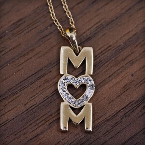 May include: A gold-colored pendant necklace with the word "MOM" spelled out. The "O" is replaced with a heart shape, encrusted with small, clear crystals. The necklace chain is also gold-colored.