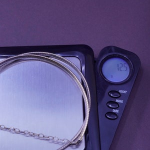May include: A silver-colored necklace with a ribbed design is placed on a digital scale. The scale displays a weight of 12.5 grams. The necklace is on a silver surface, and the scale is black.