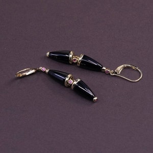 May include: A pair of gold-toned drop earrings with black, cone-shaped onyx stones. The earrings feature small pink gemstones and lever-back closures. The design is elegant and suitable for formal occasions.