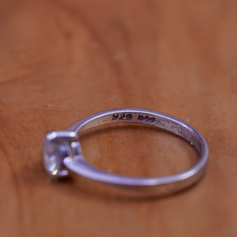 May include: A silver ring with a clear, square-cut gemstone. The ring band is stamped with "925". The ring is set against a warm, brown background. The ring is a piece of jewelry.