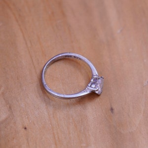 May include: A silver ring with a clear, square-cut gemstone. The ring is set on a light brown wooden surface. The band of the ring has some wear and tear. The ring is a classic design.