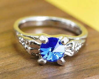 May include: Silver ring with a large, faceted blue gemstone. The ring has a detailed, textured band with decorative elements. The gemstone is held by four prongs. A close-up shot on a wooden surface.