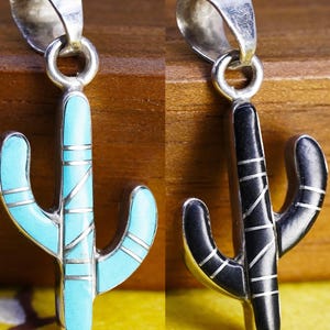 May include: Two silver pendants shaped like saguaro cacti. One pendant features inlaid turquoise stone segments, while the other features inlaid black stone segments. Both pendants have thin silver horizontal and diagonal lines across the surface.
