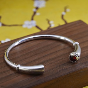 May include: A silver-colored cuff bracelet with a red gemstone set in a bezel. The bracelet has a smooth, polished finish and a simple, elegant design. The gemstone is a deep red color, and the bracelet is displayed on a wooden surface.