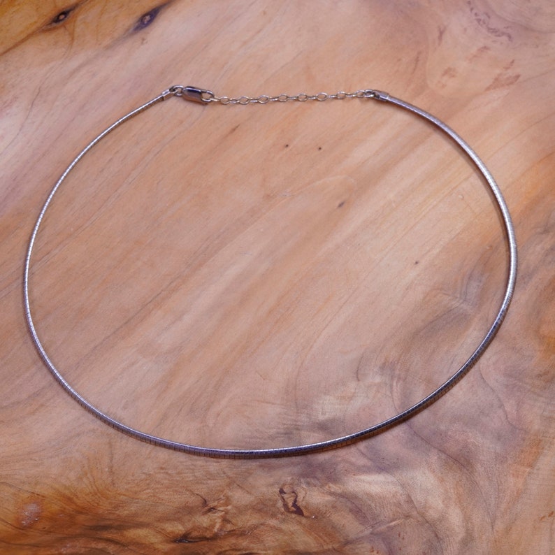 May include: A silver-colored choker necklace with a simple, elegant design. The necklace is a thin, curved band that sits close to the neck, with a small chain for adjusting the length. The clasp is visible. The necklace is displayed on a wooden surface.