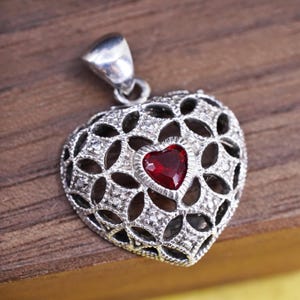 May include: Silver heart-shaped pendant with a filigree design and a small, red heart-shaped gemstone in the center. The pendant has a bail for attaching to a chain. The pendant is displayed on a wooden surface.
