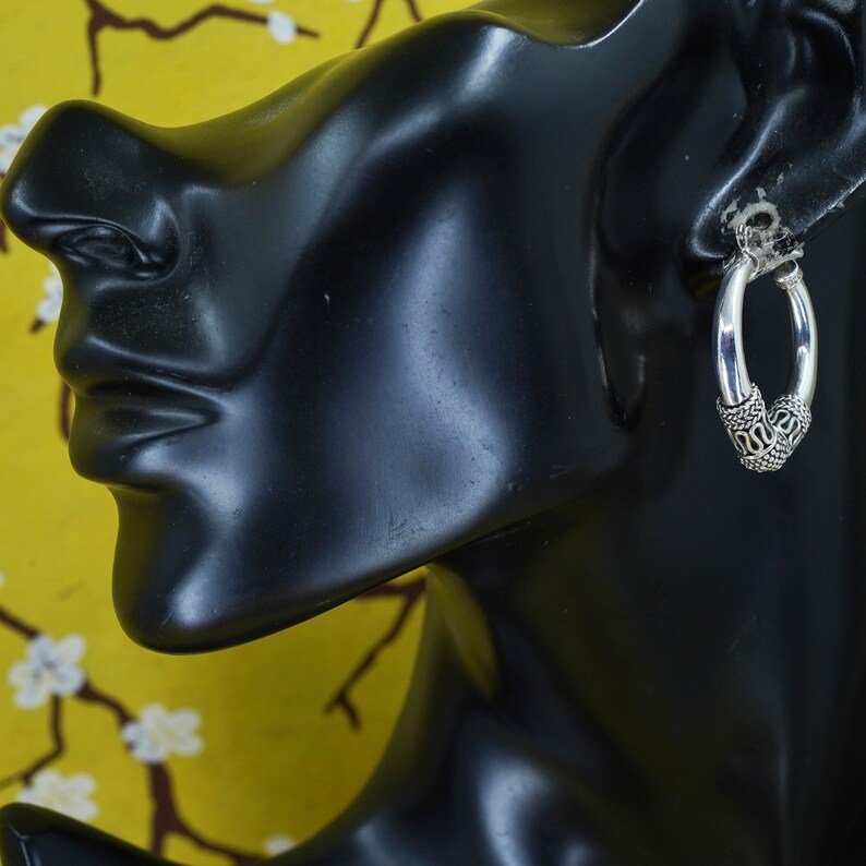 May include: Silver hoop earrings with detailed, patterned accents. The earrings are oval-shaped and feature a decorative design at the bottom. The earrings are displayed on a black mannequin head against a yellow floral background.