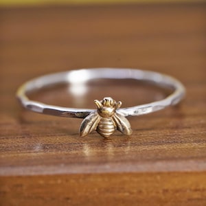 May include: A thin, silver-toned ring featuring a small, gold-toned bee charm centered on the band. The silver band has a hammered, textured finish, while the bee charm displays detailed wings and a segmented body design.