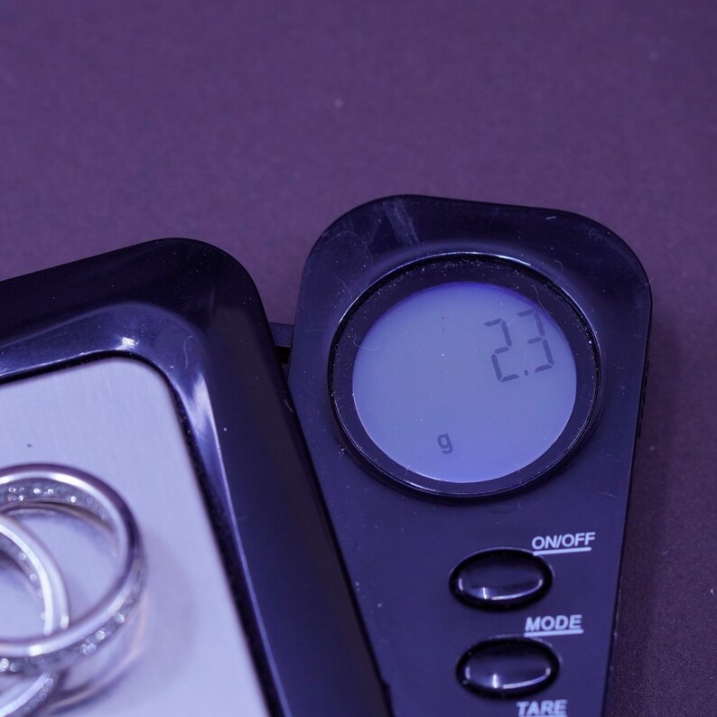 Pu&ograve; includere: Una bilancia digitale mostra 2,3 grammi. Un anello d'argento con piccole pietre trasparenti &egrave; sulla bilancia. La bilancia &egrave; nera e argentata, con un pulsante di accensione/spegnimento e altri pulsanti funzione. Lo sfondo &egrave; viola.