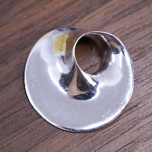 May include: A polished silver-toned pendant featuring a smooth, circular, and concave form with an off-center, funnel-like opening. The metallic surface is reflective, showing light highlights and subtle curves across its rounded, disc-shaped body.