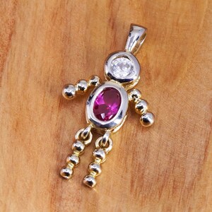 Peut inclure: Pendentif en forme de personne, avec un design argent et or. La tête est ornée d'une pierre ronde et transparente, et le corps d'une pierre ovale magenta. Les bras et les jambes sont faits de sphères dorées.
