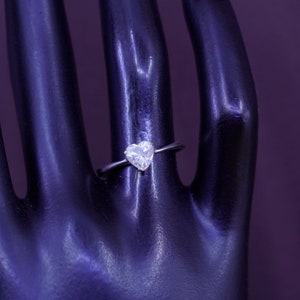 May include: A silver ring with a heart-shaped, clear gemstone. The ring is set on a thin band. The gemstone is faceted, reflecting light. The ring is displayed on a black hand against a purple background. Jewelry.