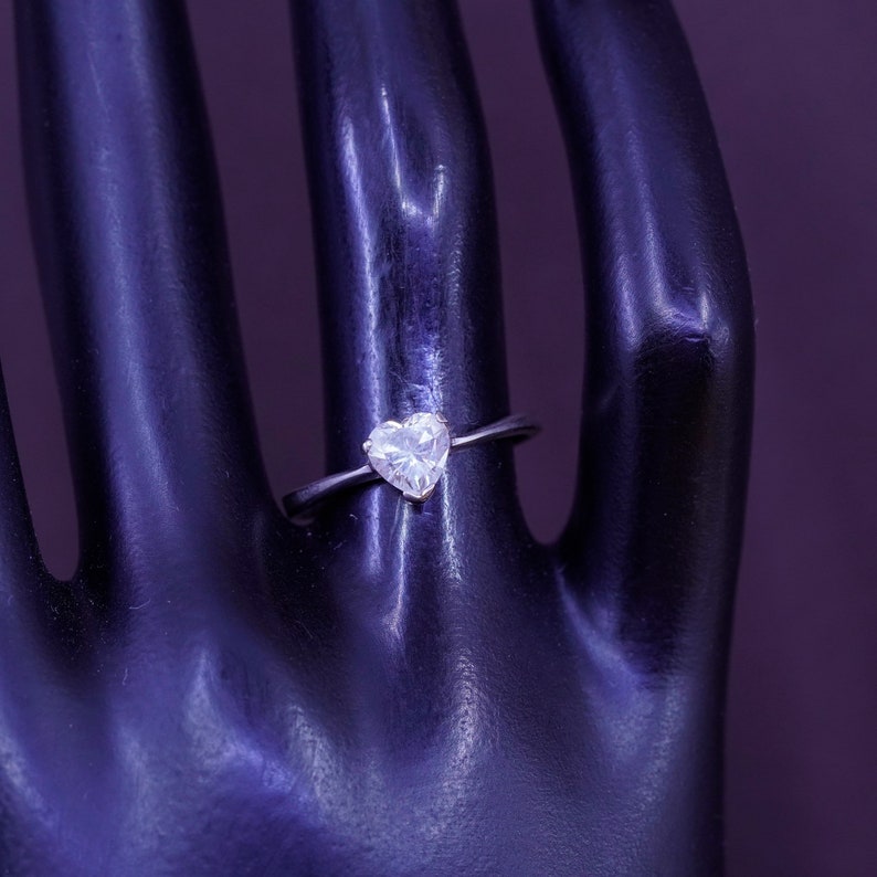 May include: A silver ring with a heart-shaped, clear gemstone. The ring is set on a thin band. The gemstone is faceted, reflecting light. The ring is displayed on a black hand against a purple background. Jewelry.