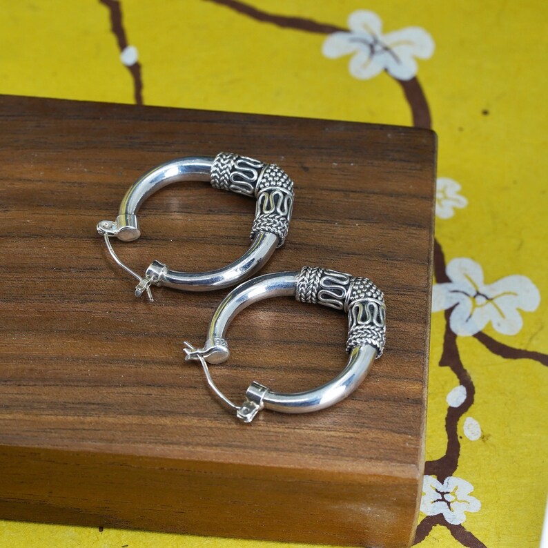 May include: A pair of silver hoop earrings with a decorative design on the top portion. The earrings are displayed on a wooden surface. The decorative design features a woven pattern with a series of curved lines. The earrings have a simple clasp closure.