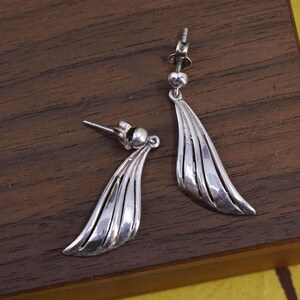 May include: A pair of silver-toned drop earrings. Each earring features a post with a small ball, connected to a stylized wing-shaped pendant with vertical grooves. The earrings are displayed on a dark wooden surface.