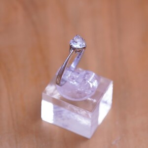 May include: A silver ring with a clear, triangular gemstone. The ring is displayed on a clear, square stand against a blurred, light brown background. The ring's band is thin and metallic.