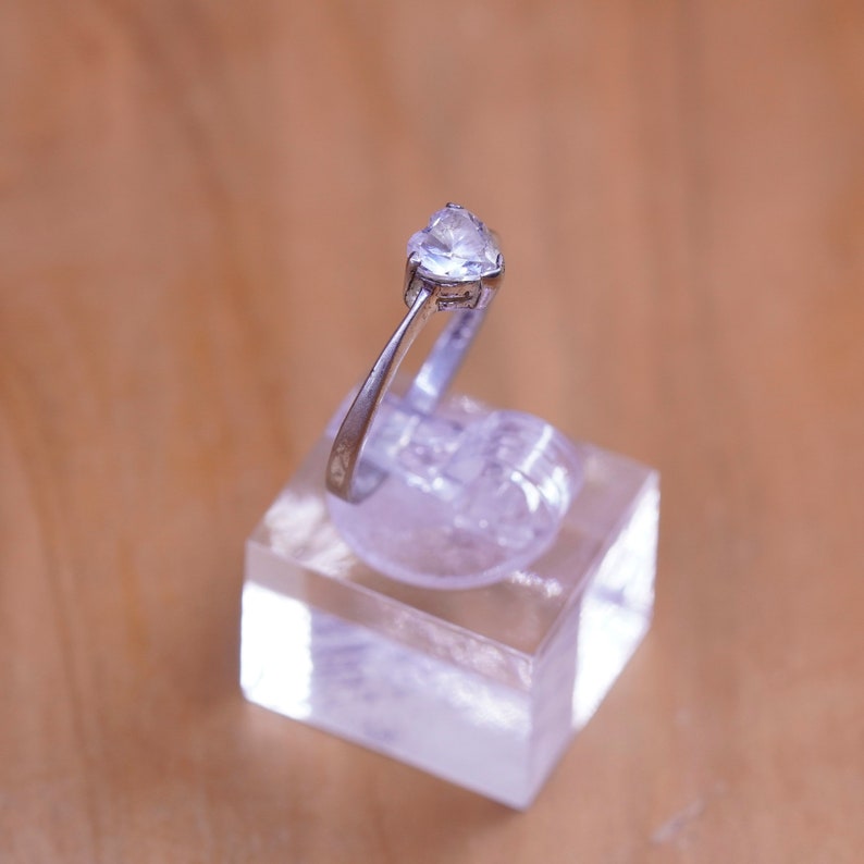 May include: A silver ring with a clear, triangular gemstone. The ring is displayed on a clear, square stand against a blurred, light brown background. The ring's band is thin and metallic.