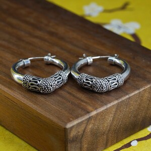 May include: A pair of silver hoop earrings. The earrings have a thick, rounded design with detailed patterns. The central section features a textured, beaded design, while the ends have a decorative, curved pattern. The earrings are displayed on a wooden surface.