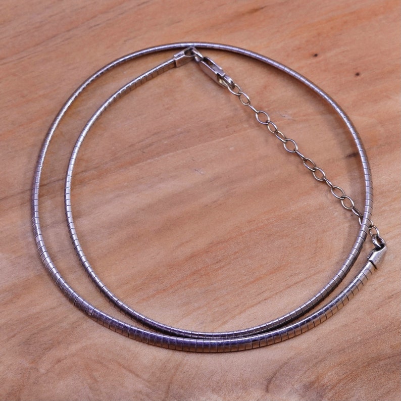 May include: A silver-toned necklace with a flexible, ribbed design. The necklace features a simple clasp and an adjustable chain. The jewelry is displayed on a light brown wooden surface.