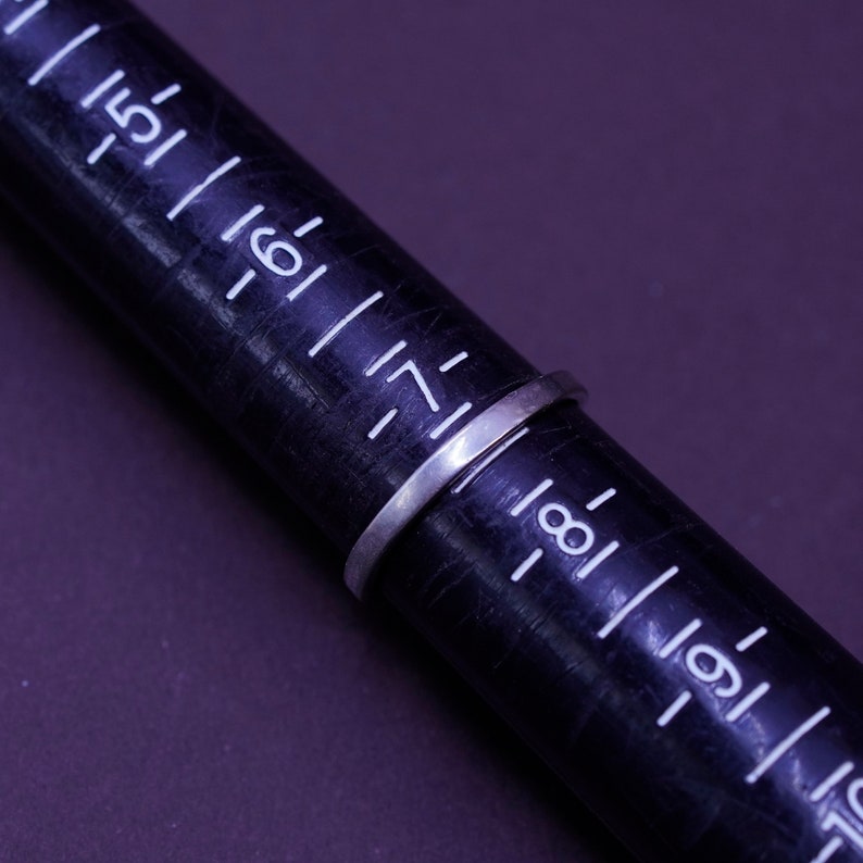 May include: A silver ring is placed on a black ring sizer. The ring is thin and appears to be made of metal. The ring sizer has white markings and numbers.