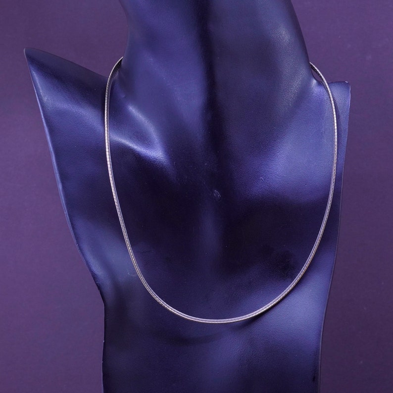 May include: A silver-toned necklace displayed on a black mannequin. The necklace is a thin, delicate chain, likely made of metal. The background is a solid purple color.