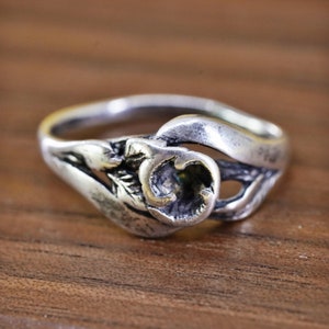May include: A silver ring with an open floral design. The ring has a detailed, textured pattern with a central setting. The band curves around the finger, with the floral elements extending outwards.