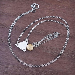 May include: A delicate silver necklace featuring a small, brushed silver triangle pendant and a textured gold-colored sphere. The necklace has a fine chain and a simple clasp. The jewelry is displayed on a wooden surface.