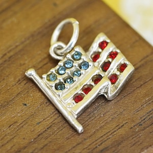 May include: A silver charm in the shape of the American flag. The flag features blue and red gemstones, representing the stars and stripes. The charm has a small loop at the top for attaching to a necklace or bracelet.