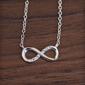 May include: A silver-colored infinity symbol pendant necklace with small clear gemstones. The necklace has a delicate chain and the infinity symbol is centered in the image. The background is a dark wood surface.