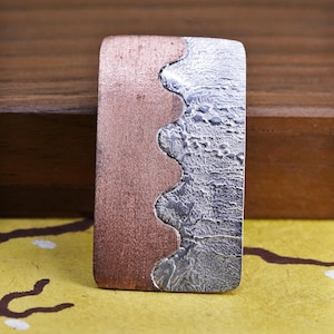 May include: A rectangular metal pendant with a two-tone design. The left side is a reddish-brown color, while the right side has a textured, silver appearance. The two sections are separated by a wavy line. The pendant is displayed on a yellow surface.