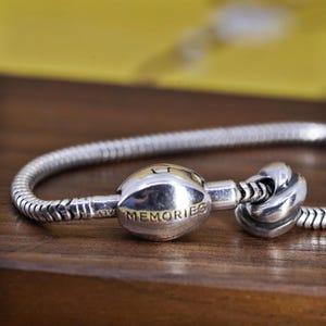 Puede incluir: Una pulsera plateada con un cierre que dice "MEMORIES" en letras doradas. La pulsera está sobre una superficie de madera. El cierre tiene forma redondeada y un diseño de nudo en el otro extremo.