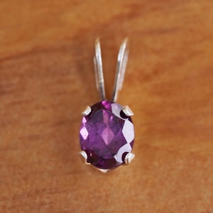 May include: A silver pendant featuring an oval, faceted, deep purple gemstone. The gemstone is set in a silver bezel with four prongs. The pendant has a simple, elegant design, suitable for everyday wear or special occasions.