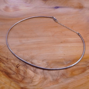 May include: A silver-colored necklace with a rounded, flexible design. The necklace has a simple, elegant style and a clasp closure. The jewelry is displayed on a light brown wooden surface.