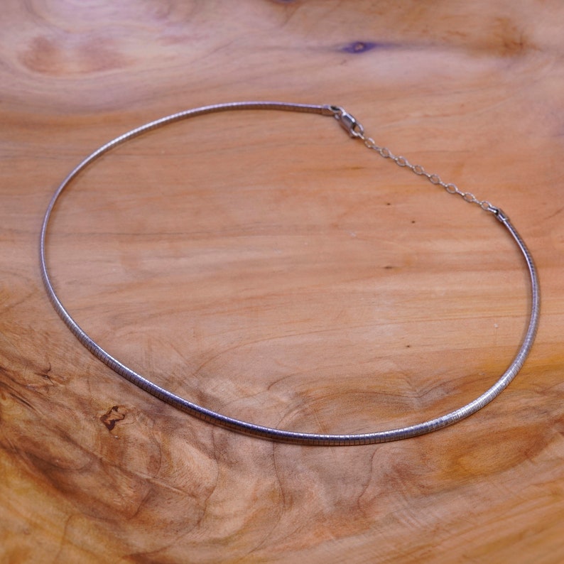 May include: A silver-colored necklace with a rounded, flexible design. The necklace has a simple, elegant style and a clasp closure. The jewelry is displayed on a light brown wooden surface.