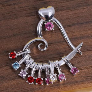May include: A silver-toned metal pendant shaped like an open heart with a small solid heart at the top. The pendant features a series of dangling charms, each set with a small round faceted gemstone in various colors including red, pink, and blue.