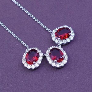 May include: A silver necklace featuring three oval-cut, deep red gemstones, each surrounded by a halo of small, clear crystals. The necklace has a delicate chain and is set against a dark purple background.