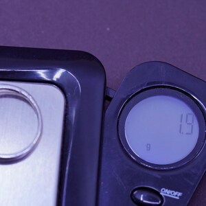 May include: A silver ring rests on a digital scale, next to the digital display showing 1.9 grams. The scale is black with a silver weighing surface. The background is a dark purple.