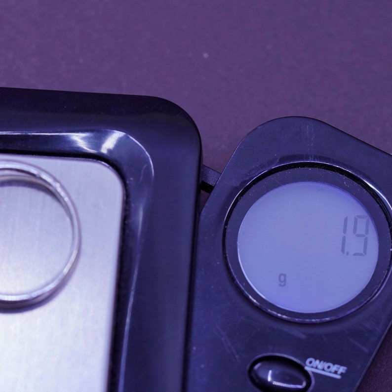 May include: A silver ring rests on a digital scale, next to the digital display showing 1.9 grams. The scale is black with a silver weighing surface. The background is a dark purple.