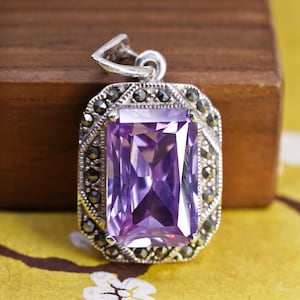 May include: A rectangular pendant featuring a large, faceted purple gemstone set in a silver-toned frame. The border is decorated with small, dark, round stones arranged in a geometric pattern, and a silver-toned bail is attached to the top.