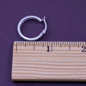Pu&ograve; includere: Un orecchino a cerchio argentato con chiusura a cerniera, incastonato con piccole pietre trasparenti. L'orecchino ha un diametro di circa 2,5 cm ed &egrave; posto su uno sfondo viola. Un righello di legno &egrave; posto accanto all'orecchino.